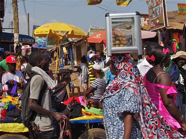 Woman carrying a plastic box of eggs on her head through a market.